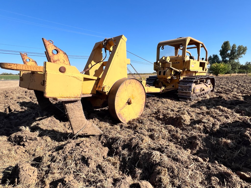 Close up view of ripper behind a crawler tractor with blue sky behind and brown dirt in foreground.