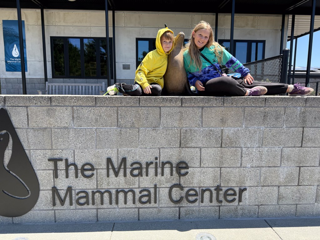 Two kids leaning on a sea lion sculpture above a sign for The Marine Mammal Center.