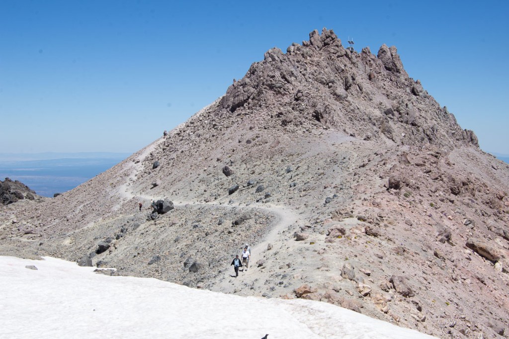 Trail across snow patch and up a rocky slope to the peak with blue sky in background.
