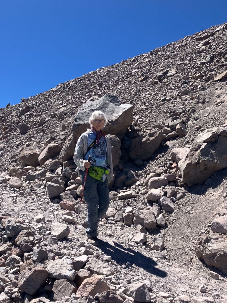 Hiker on trail with rocky slope and blue sky behind.
