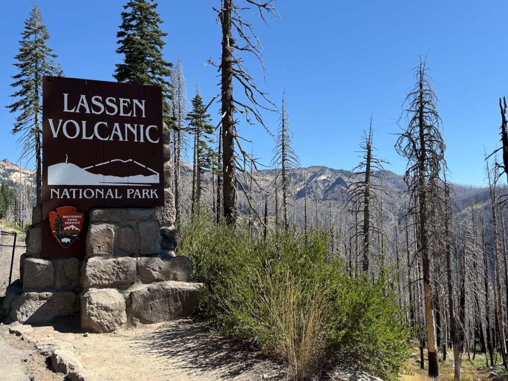Wooden sign that says Lassen Volcanic National Park with burned landscape behind.
