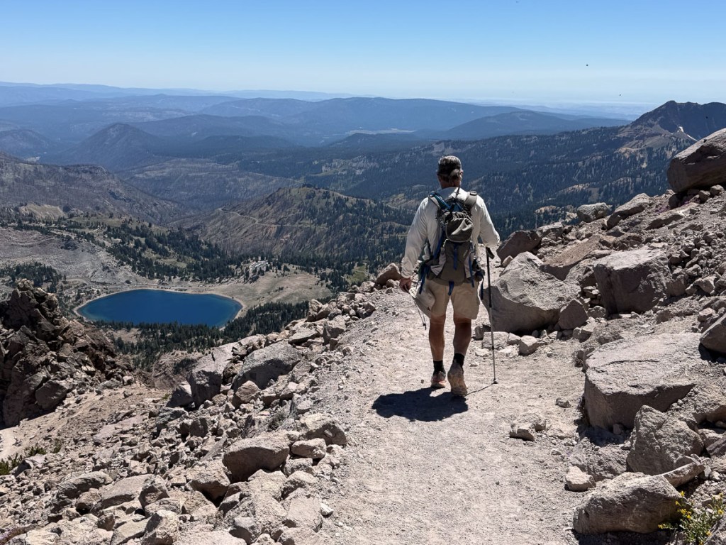 Hiker on rocky trail with blue lake and hills in distance. 