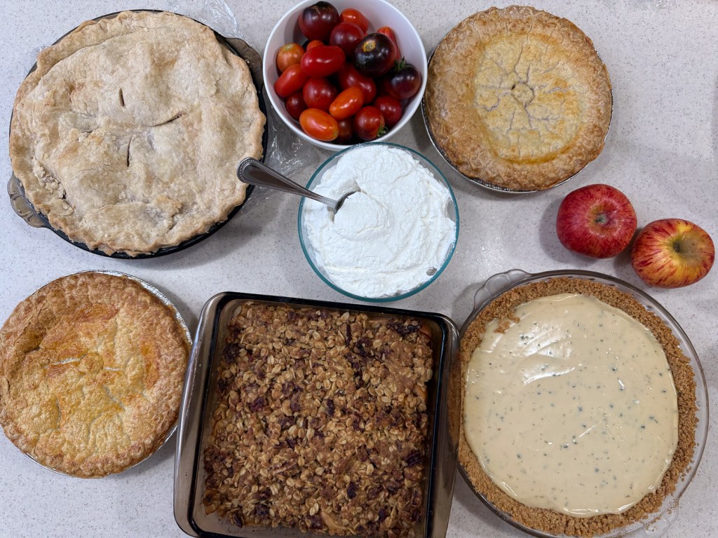 Four pies, an apple crisp, bowl of whipped cream, and tomatoes.