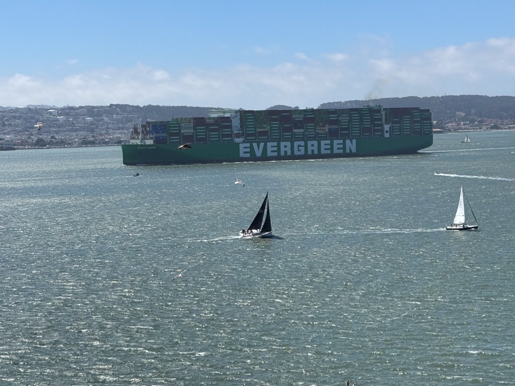 Large container ship, labeled Evergreen, in the bay with sailboats in the foreground and San Francisco hills in background.
