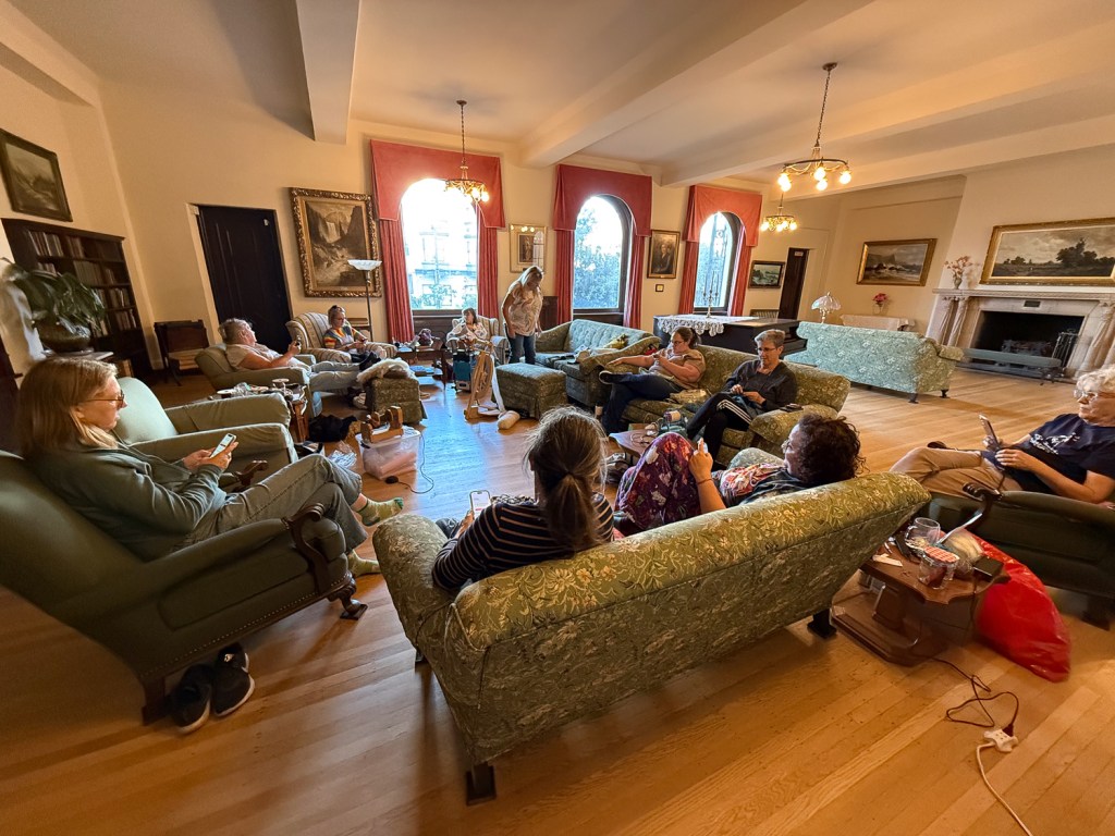 People seated on couches and chairs arranged in a circle in large room with wooden floor and arched windows. 